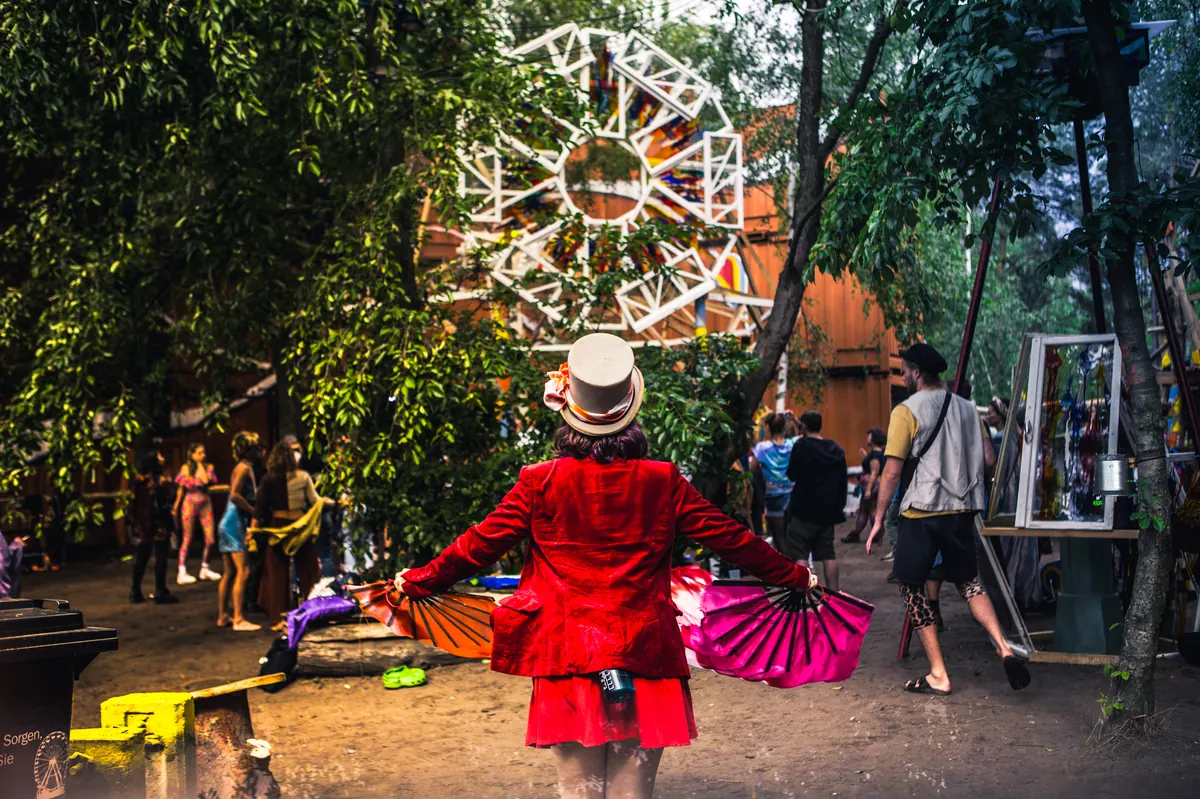 A person in a red jacket and top hat seen from behind, holding two folding fans on a path between trees